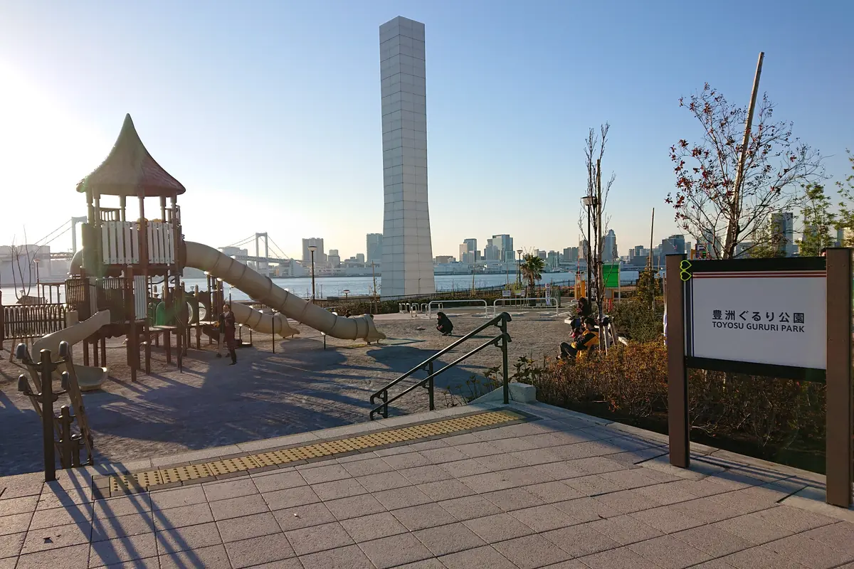 Un parque infantil con un tobogán y una alta torre de tonos claros en el parque Toyosu Gururi Park bajo la luz baja del atardecer, con la bahía de Tokio y el Rainbow Bridge al fondo y un cartel del parque a la derecha