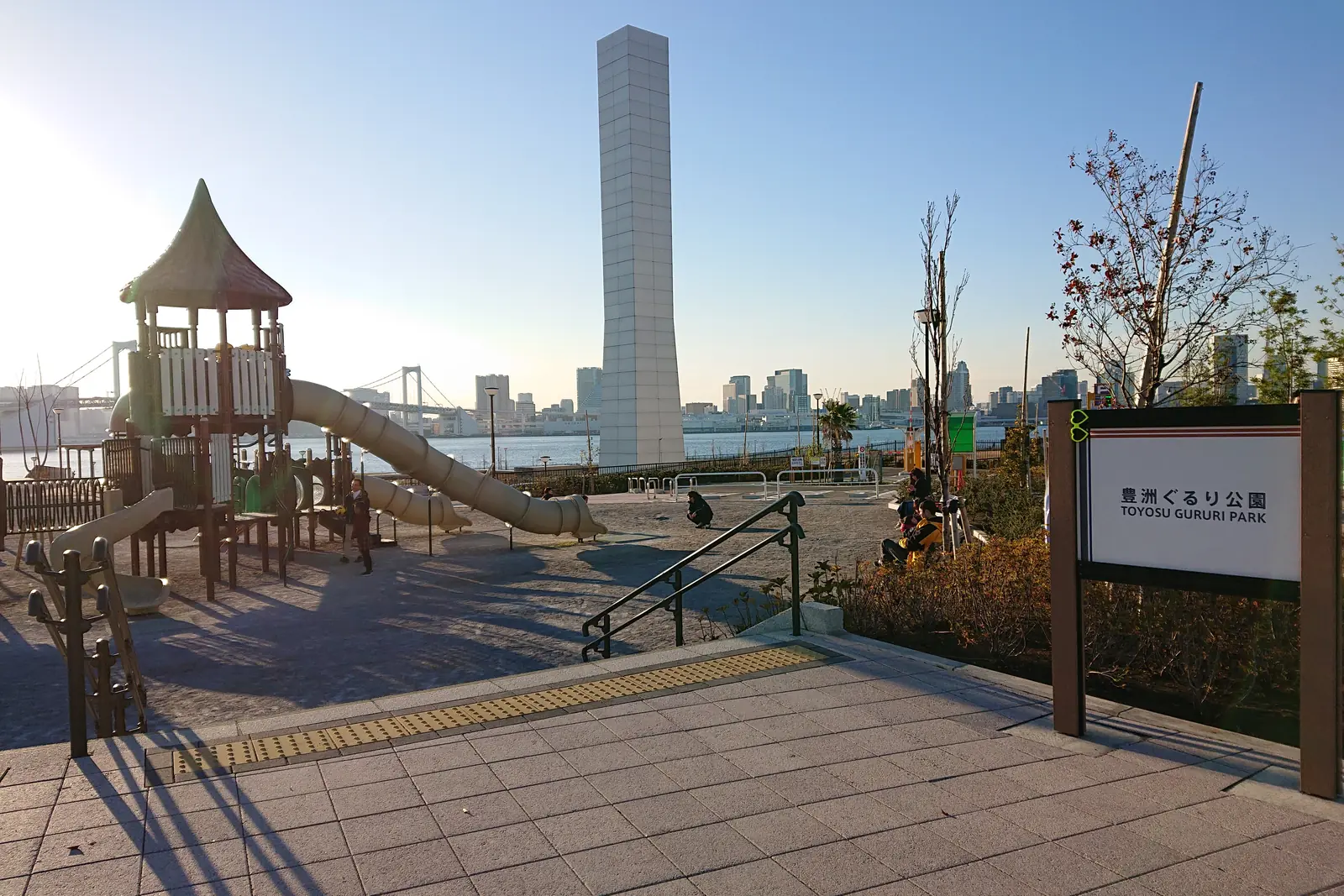 Toyosu Gururi Park'ta alçak güneş ışığında kaydıraklı bir oyun alanı ve yüksek, soluk bir kule; ötesinde Tokyo Bay ve Rainbow Bridge; müzenin değil, Toyosu sahil şeridinin genel bir fotoğrafı