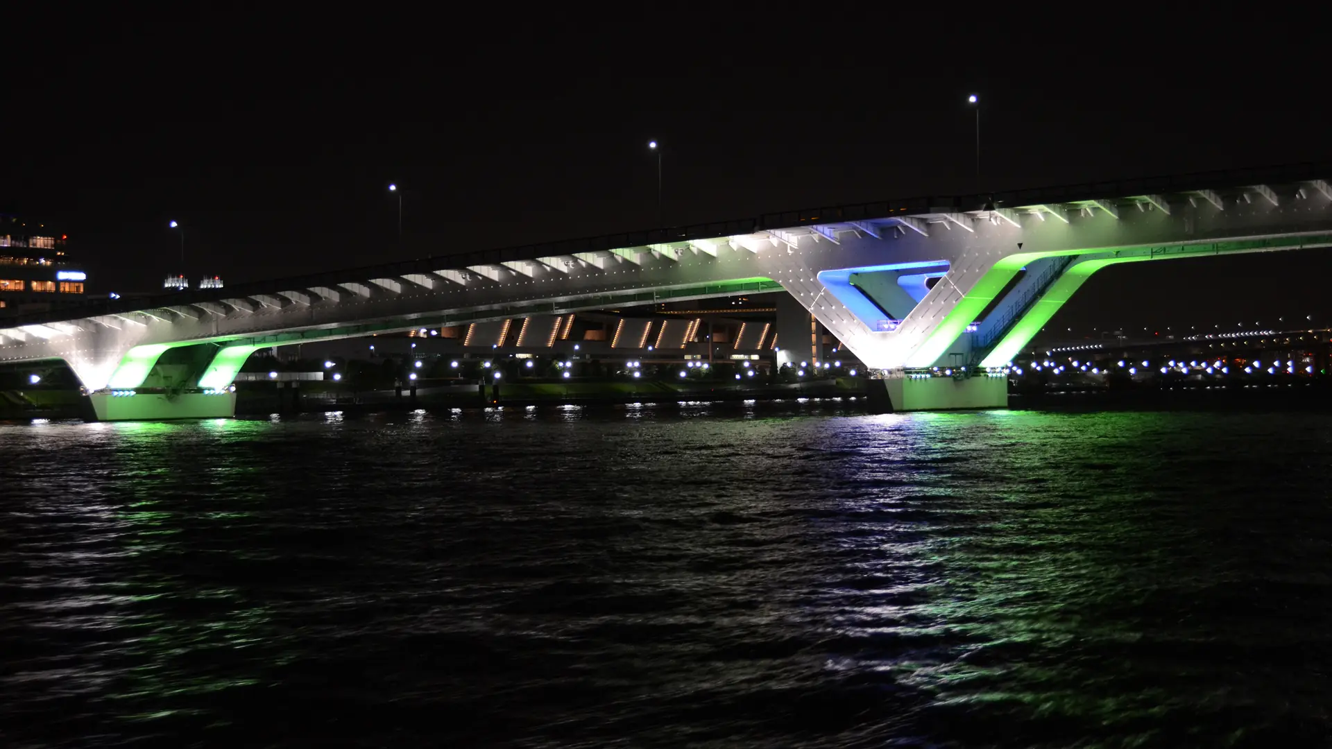 Gece karanlık suyun üzerinde yeşil, mavi ve beyaz ışıklarla aydınlanan Toyosu Ōhashi köprüsü ve ışıkların dalgalarda yansıması; teamLab Planets veya eserlerine değil, Toyosu sahil şeridine ait genel bir fotoğraf