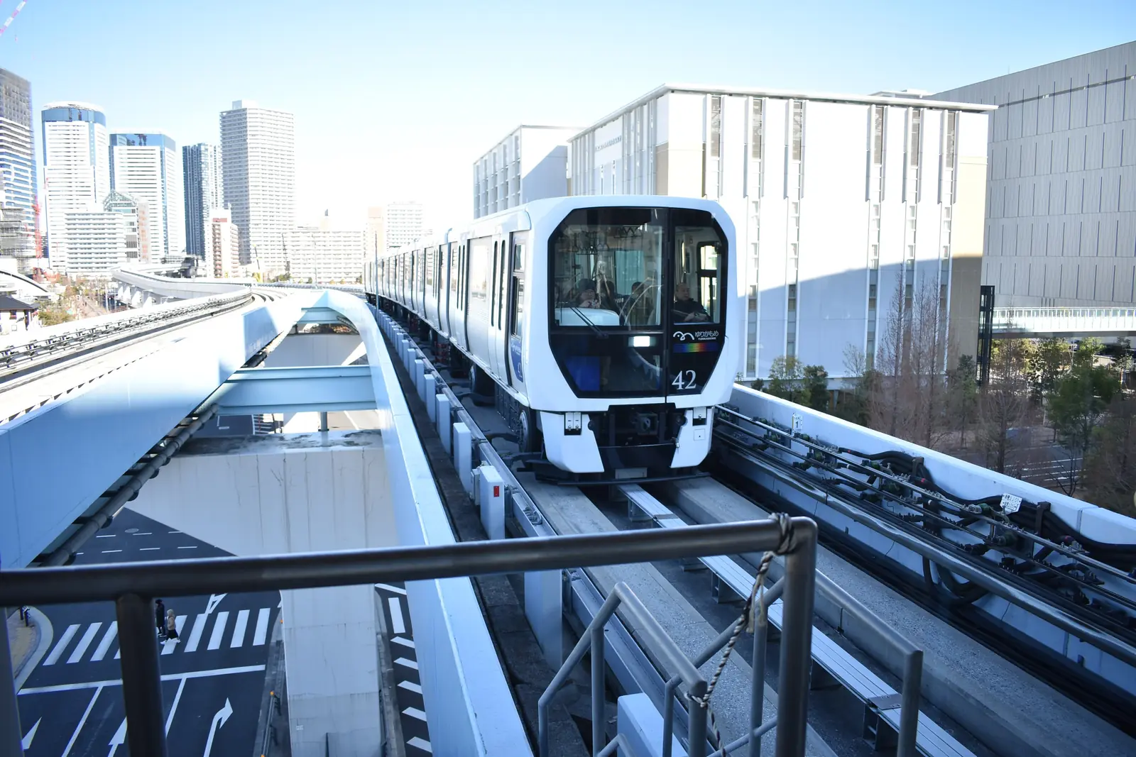 Shin-Toyosu İstasyonu'nda, cadde kavşağının üzerindeki yükseltilmiş rayında ilerleyen beyaz bir Yurikamome treni ve arkasında cam kuleler
